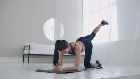 Home-exercises-for-a-beautiful-body.-a-slender-brunette-in-sportswear-is-lifting-her-legs-back-while-standing-on-all-fours-on-the-rug